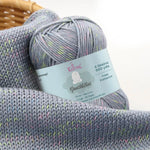 KnitPal GentleBliss DK weight yarn in soft silver gray with multicolor marl flecks, skein on knitted fabric swatch, label visible