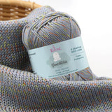 KnitPal GentleBliss DK weight yarn in medium gray with orange and yellow-green marl flecks, skein on knitted fabric swatch, label visible