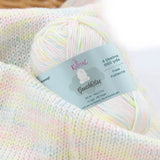 KnitPal GentleBliss DK weight yarn in white with soft pastel pink, yellow, and blue marl flecks, skein on knitted fabric swatch, label visible