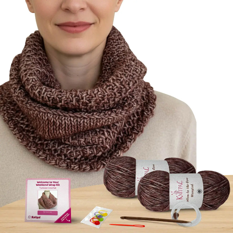 Woman wearing a brown knitted scarf with yarn balls and a kit on a wooden surface.