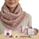 Person wearing a pink knitted scarf with yarn balls and knitting tools on a wooden surface.