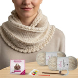 Person wearing a beige knitted cowl with yarn, needles, and packaging on a wooden surface.